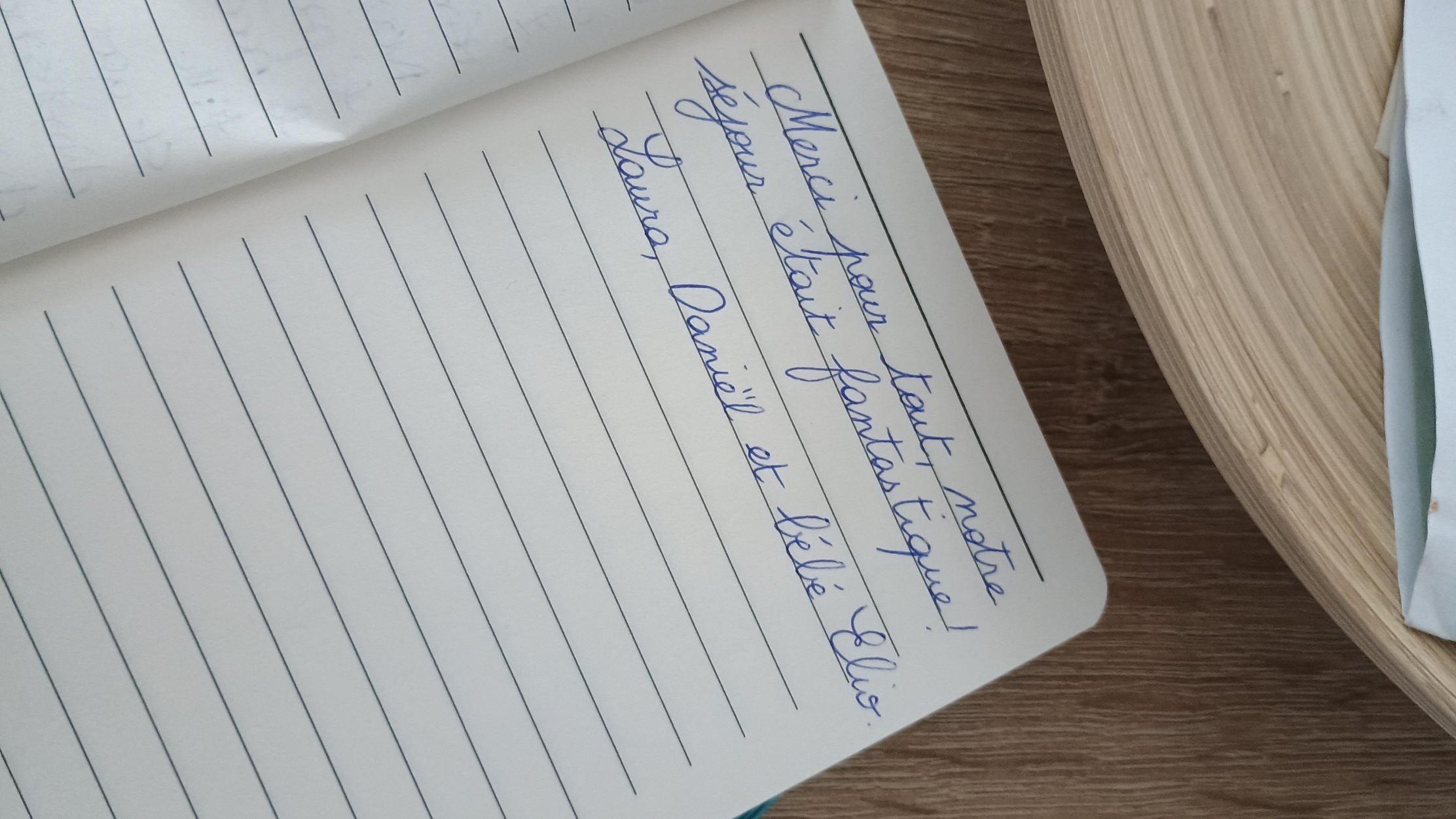 Un cahier ligné ouvert montrant une écriture manuscrite en français bleu, qui dit : "Merci pour tout, notre séjour était fantastique ! Laura, Daniel et bébé Elise."