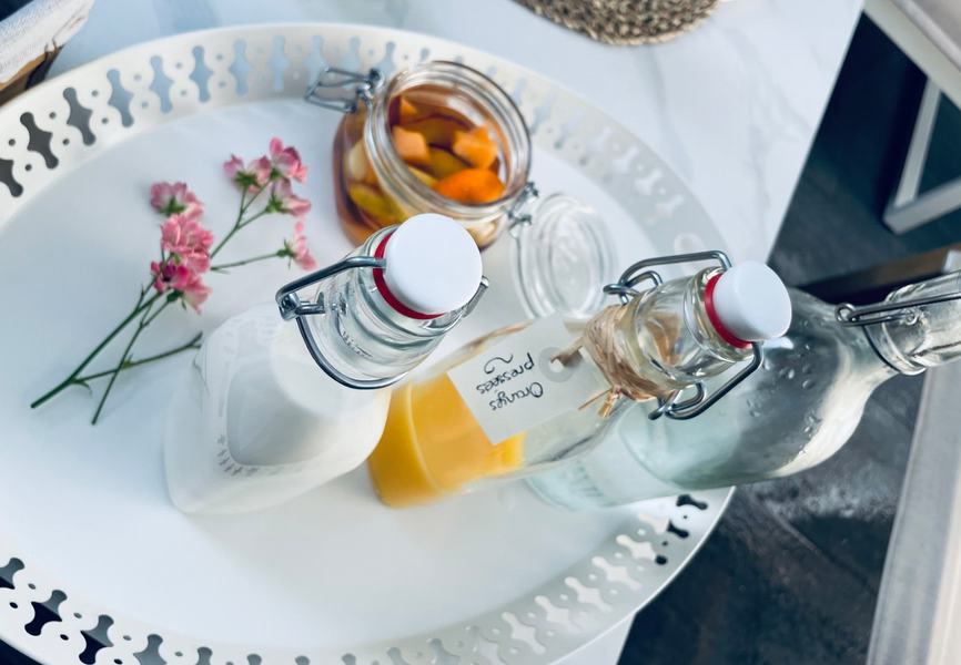 Un plateau blanc ajouré contenant plusieurs petites bouteilles en verre avec bouchon mécanique, l'une remplie de lait, une autre de jus d'orange étiquetée "Oranges pressées", et un pot de fruits frais coupés, avec quelques fleurs roses.