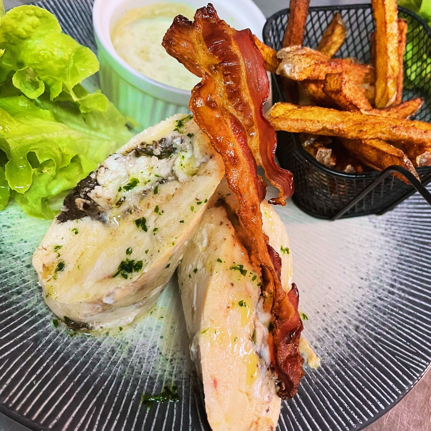 Un plat principal composé d'une poitrine de poulet farcie, de deux tranches de bacon croustillant, d'une salade verte et d'une portion de frites.