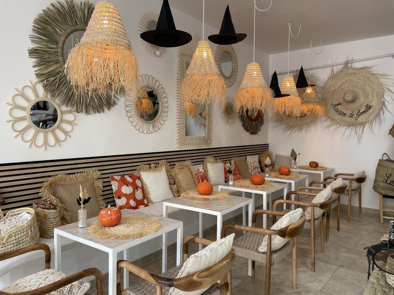La photo montre l’espace salon, aménagé avec plusieurs tables et fauteuils en bois. Les miroirs, suspensions en fibres végétales et décorations murales composent une ambiance bohème et accueillante. Quelques éléments saisonniers viennent compléter la décoration.