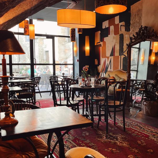 L'intérieur d'un restaurant ou d'un café, avec des tables et des chaises en bois sombre sur un tapis persan rouge, des luminaires suspendus jaunes, un miroir ancien, et une grande fenêtre donnant sur l'extérieur.