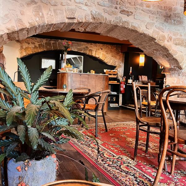 L'intérieur d'un restaurant ou d'un café, avec des murs en pierre, des arches, un tapis persan rouge, des tables et chaises en bois, et une plante verte au premier plan.
