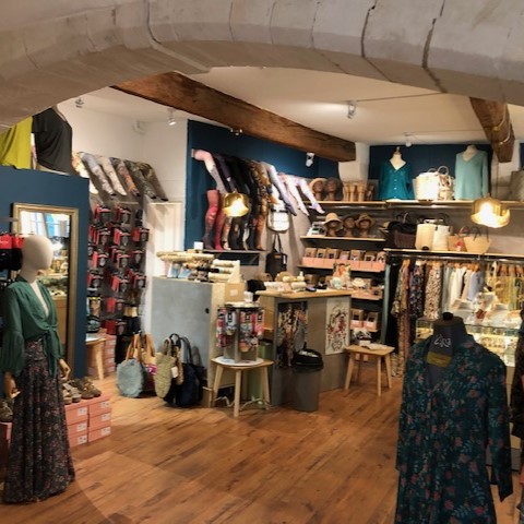 L'intérieur d'une boutique de vêtements et d'accessoires, avec un sol en parquet, des étagères et des portants remplis d'articles, et des mannequins présentant des tenues, sous un plafond avec des poutres en bois.