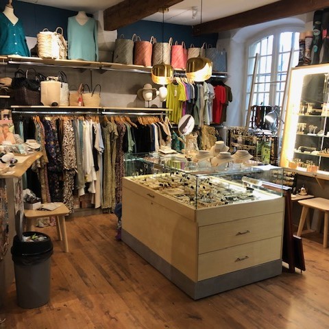 L'intérieur d'une boutique de mode, avec un sol en parquet, des vêtements suspendus sur des portants, des sacs et accessoires sur des étagères, et une grande vitrine remplie de bijoux au centre.