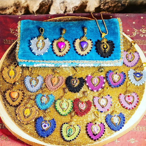 Une collection de pendentifs en forme de cœur, richement brodés de perles colorées et de pierres, disposés sur un plateau circulaire doré et un petit sac pochette en velours turquoise.