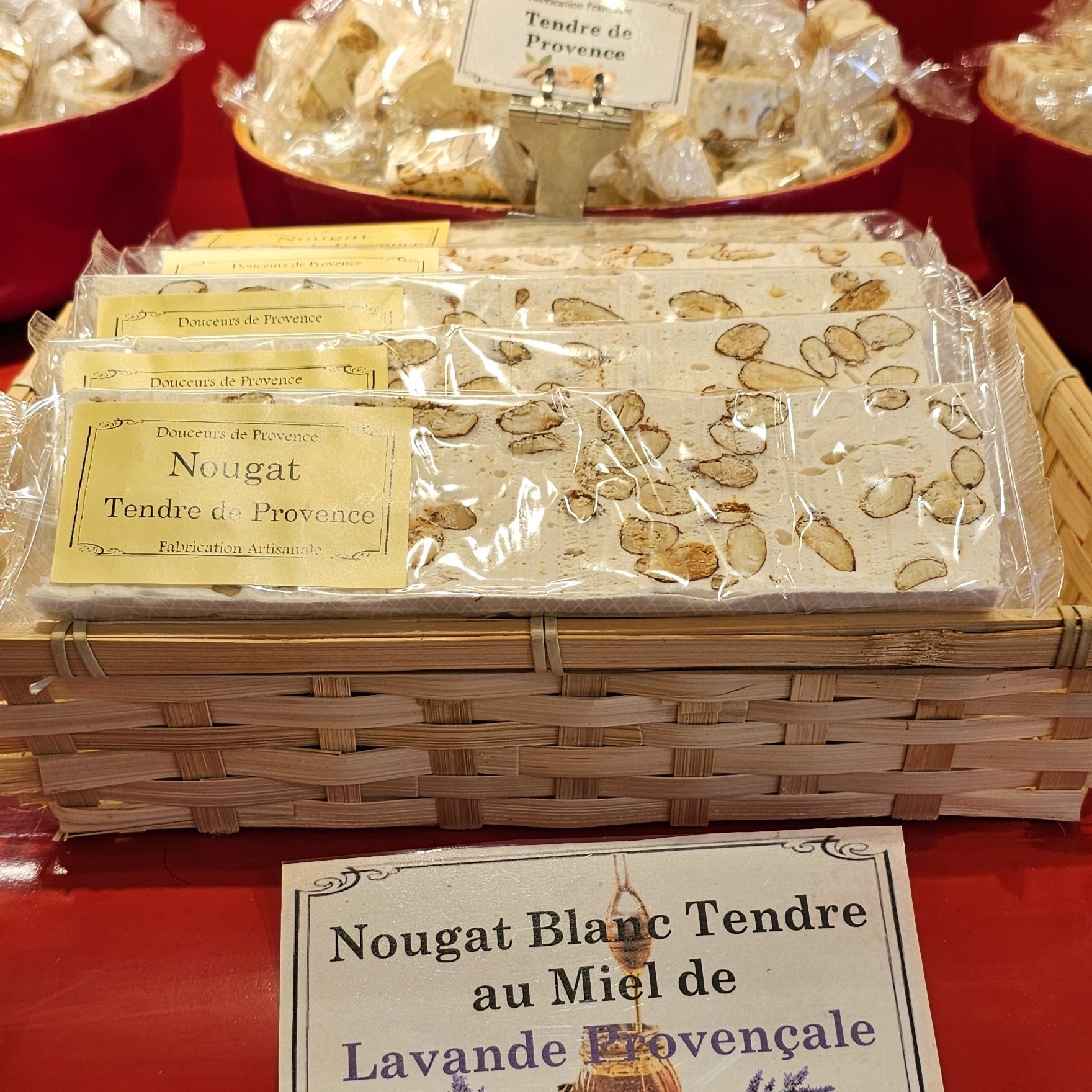 Un plateau en osier rempli de paquets individuels de nougat blanc tendre aux amandes, étiquetés "Nougat Tendre de Provence, Fabrication Artisanale". Un panneau devant le plateau indique "Nougat Blanc Tendre au Miel de Lavande Provençale".